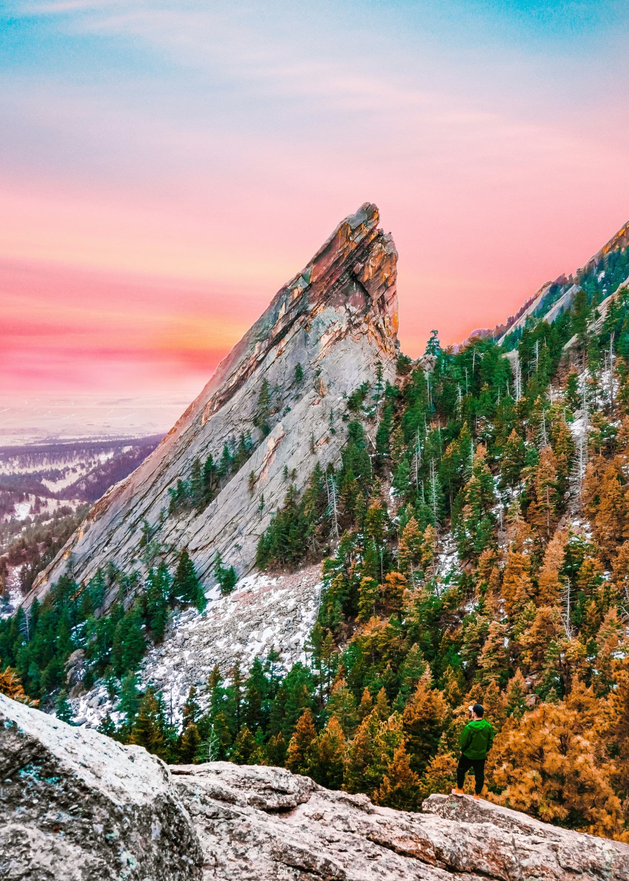 A person at the base of a steep, forested mountain peak, gazing up towards the vibrant sunset sky, in October in Aspen.