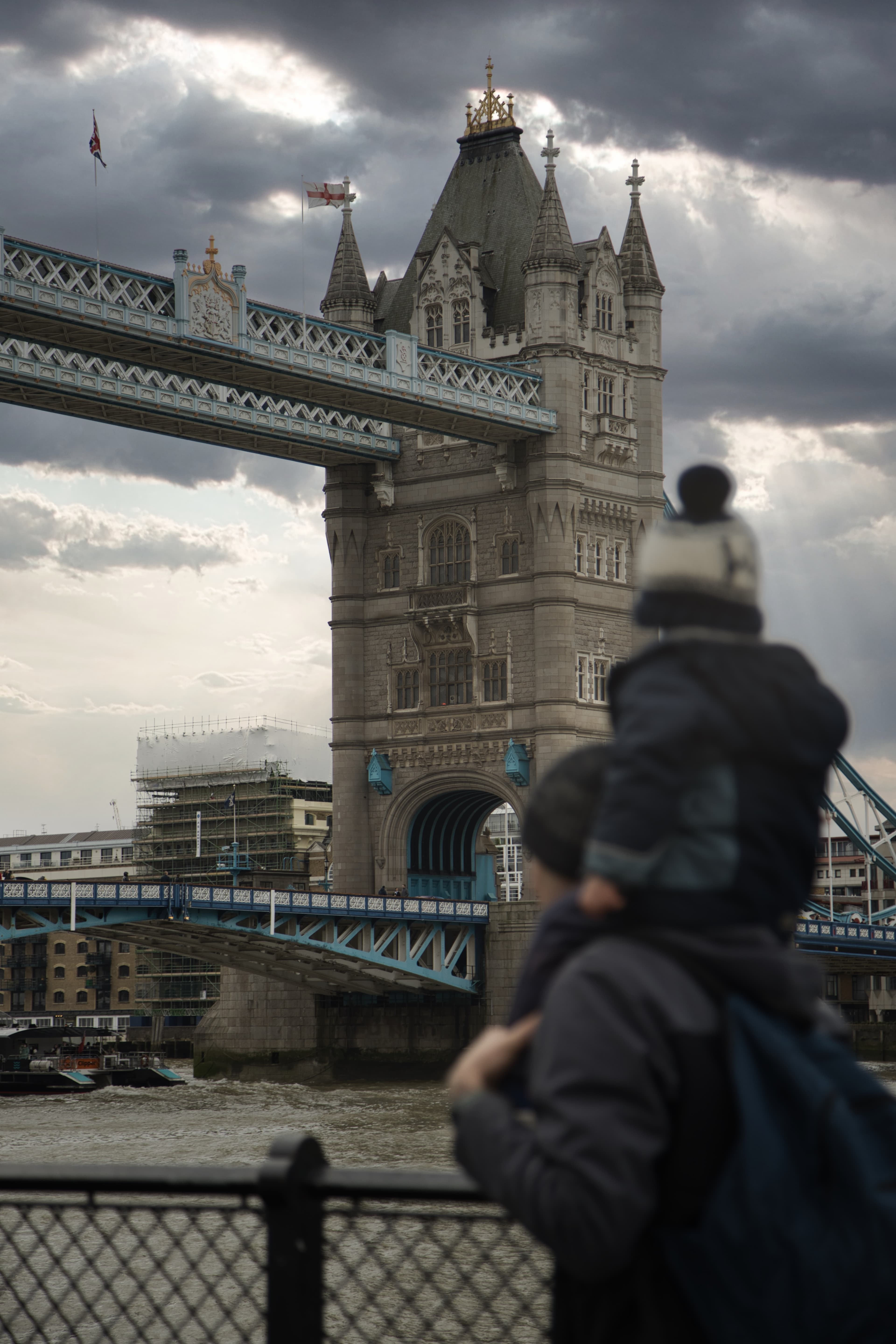 Man with child on shoulders walks past bridge and building on cloudy day