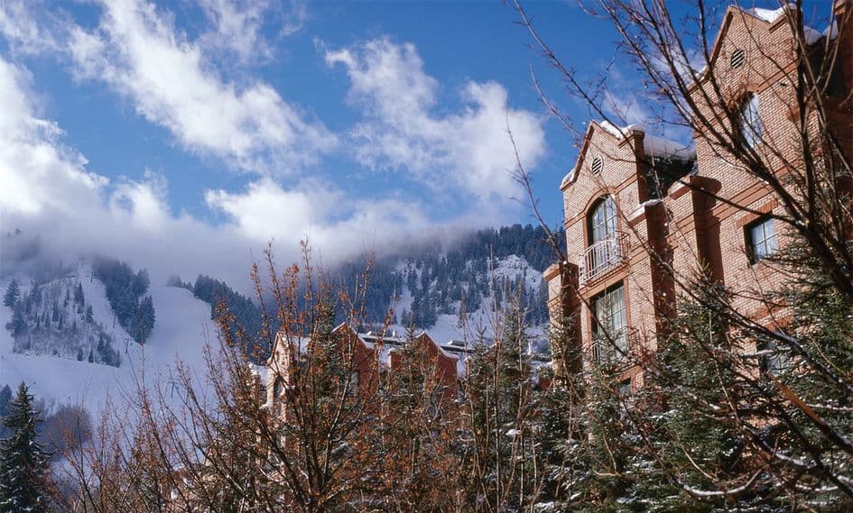 The St. Regis Hotel in Aspen reaches to the sky and overlooks a mountainous range on a sunny day with wisps of clouds cascading down the hills.