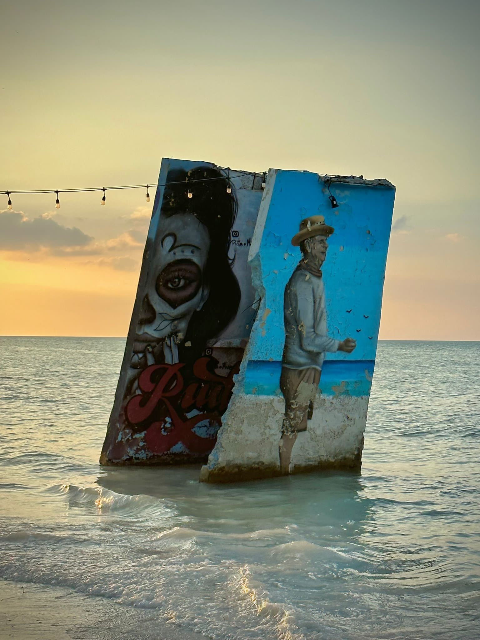 A picture of a mural painted on a large rock positioned at an angle in the ocean water. There are string lights tied to the left side of the rock, and a vibrant orange sunset in the background.
