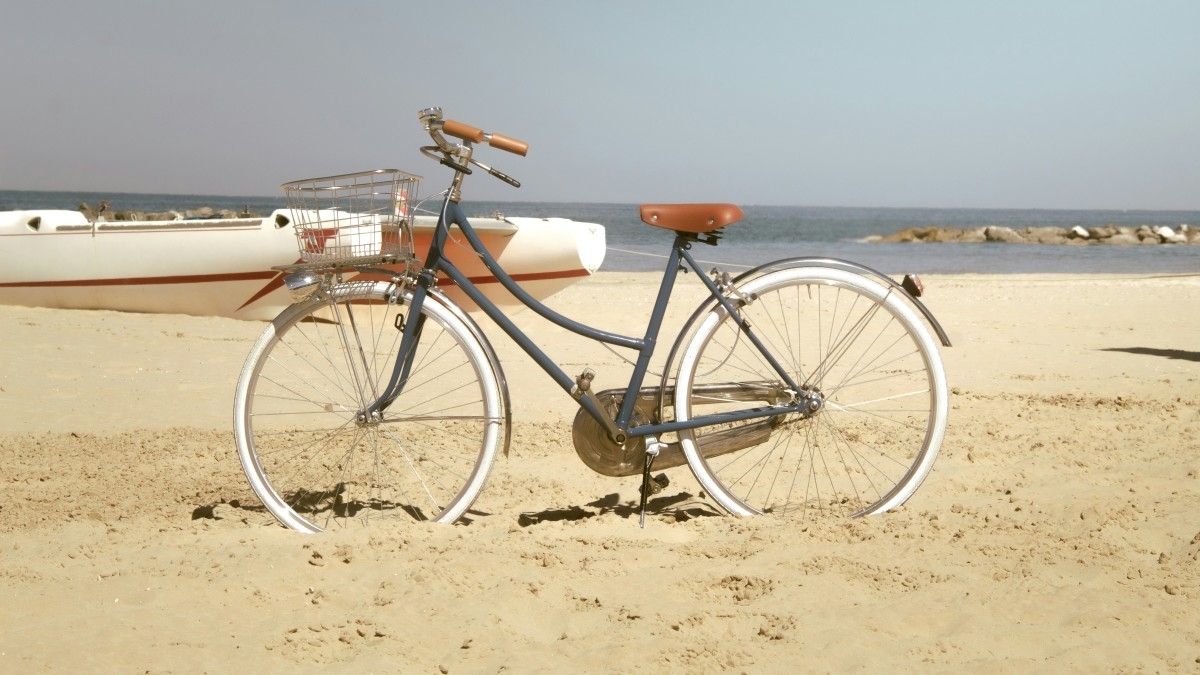 A beach cruiser bicycle parked on the beach with boats and a harbor in the background.