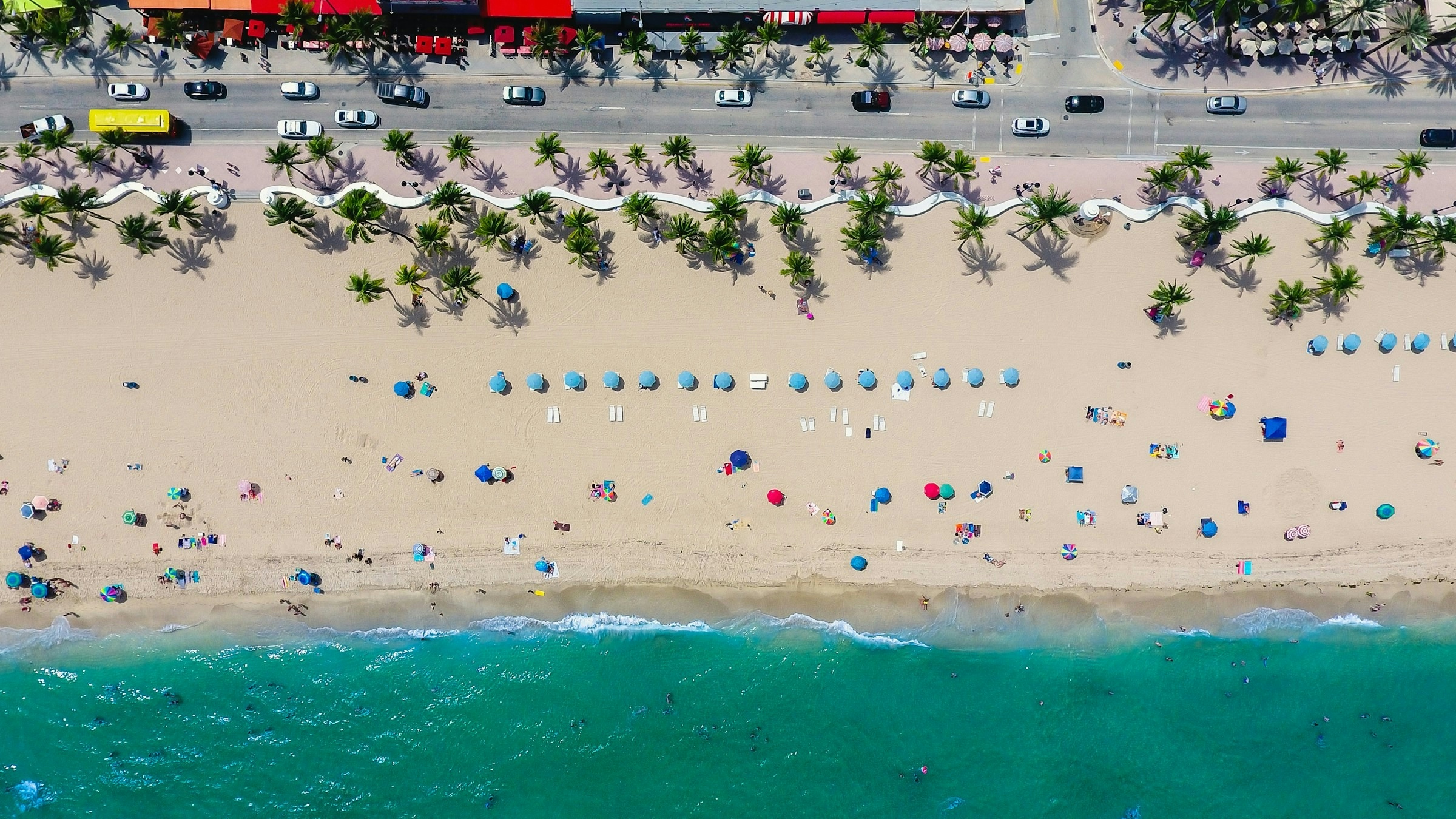 Birds-eye view of a beach, ocean, and busy street with cars. The beach has different colored umbrellas and palm trees.