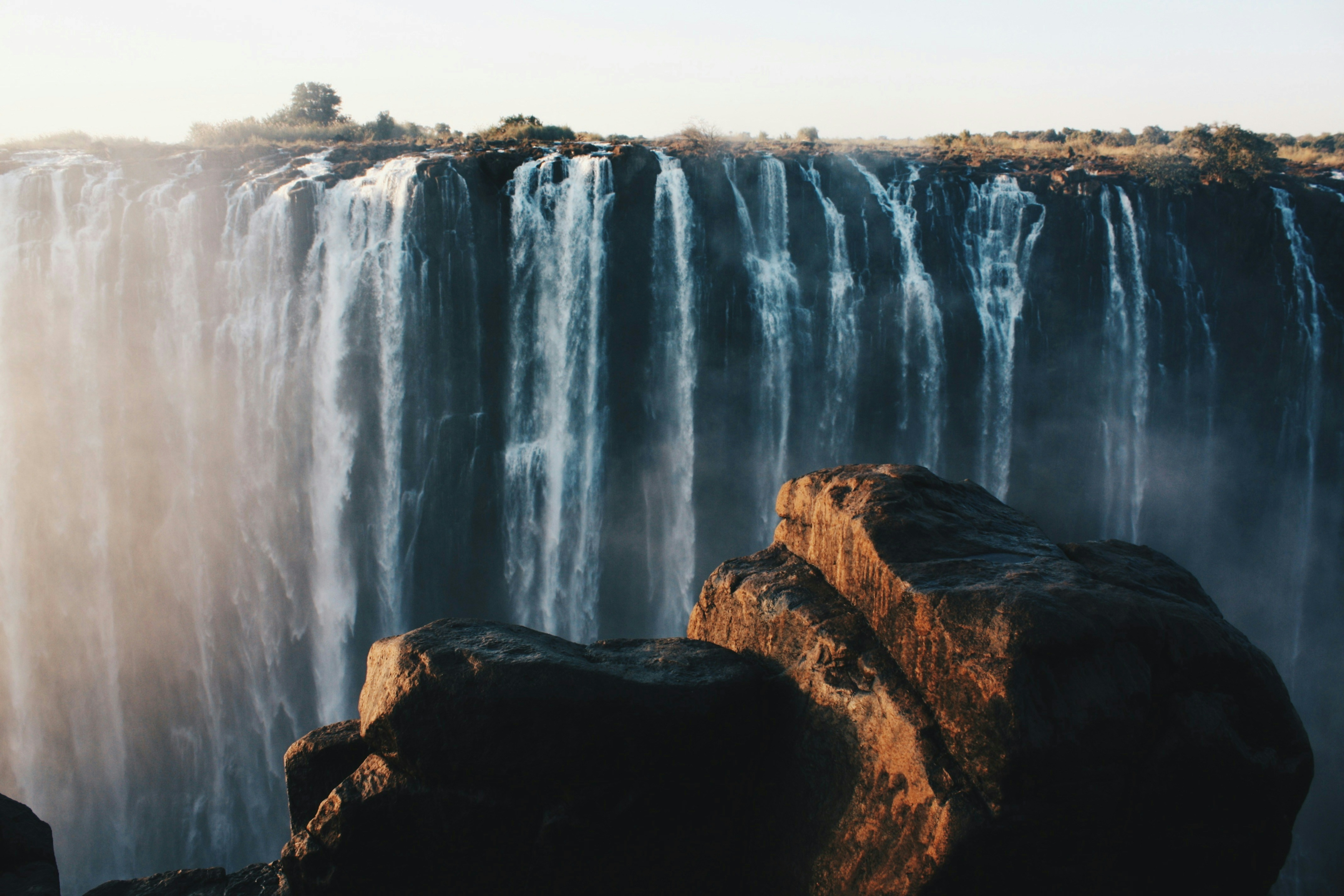 Victoria Falls in Zimbabwe, large waterfalls as seen across a canyon with rocks in the foreground.