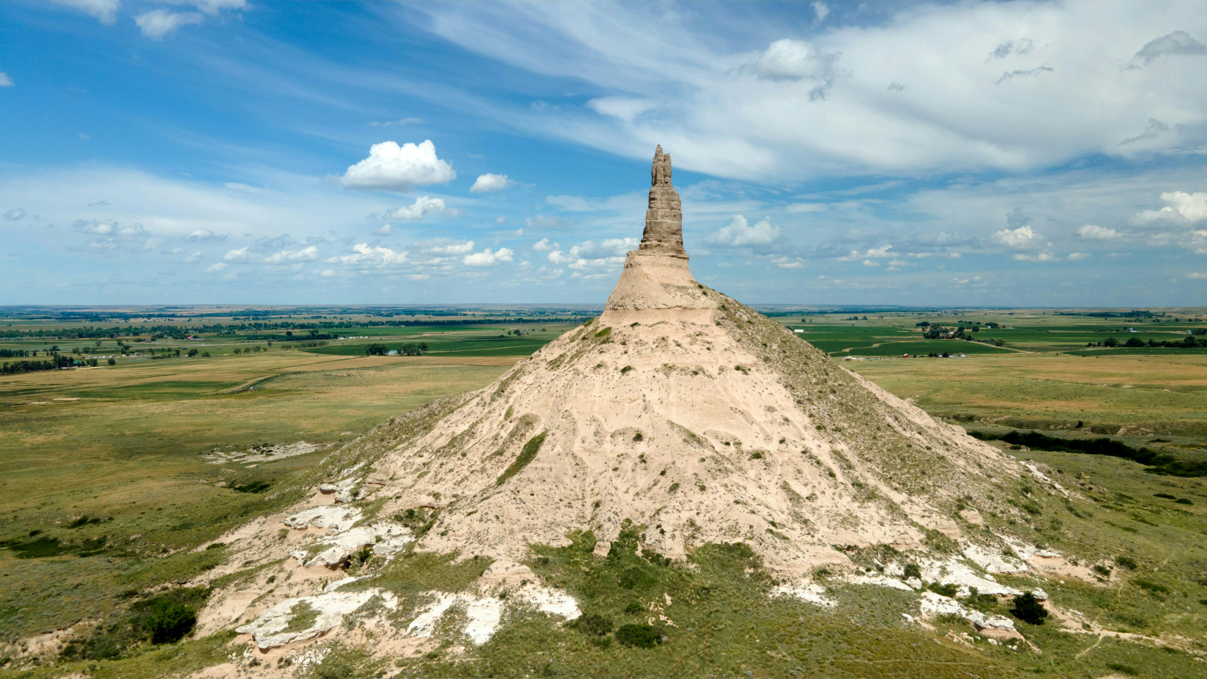 Chimney Rock, a large pointed rock formation in the middle of a green plain with blue sky.