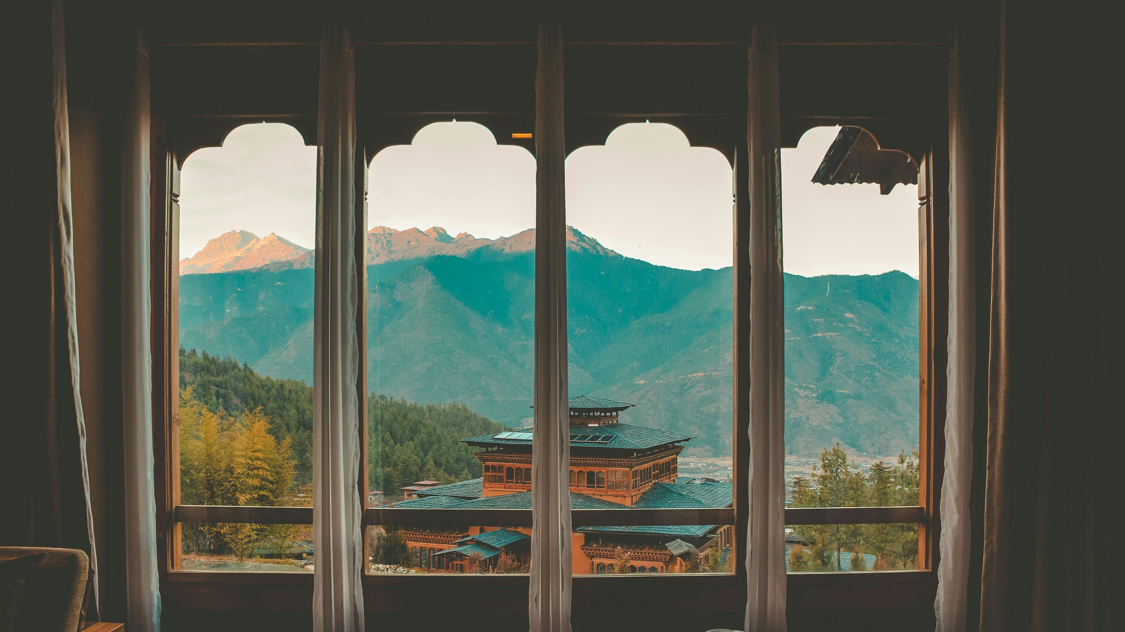A hotel room with white curtains and wood floors looking out at a traditional building in front of mountains.