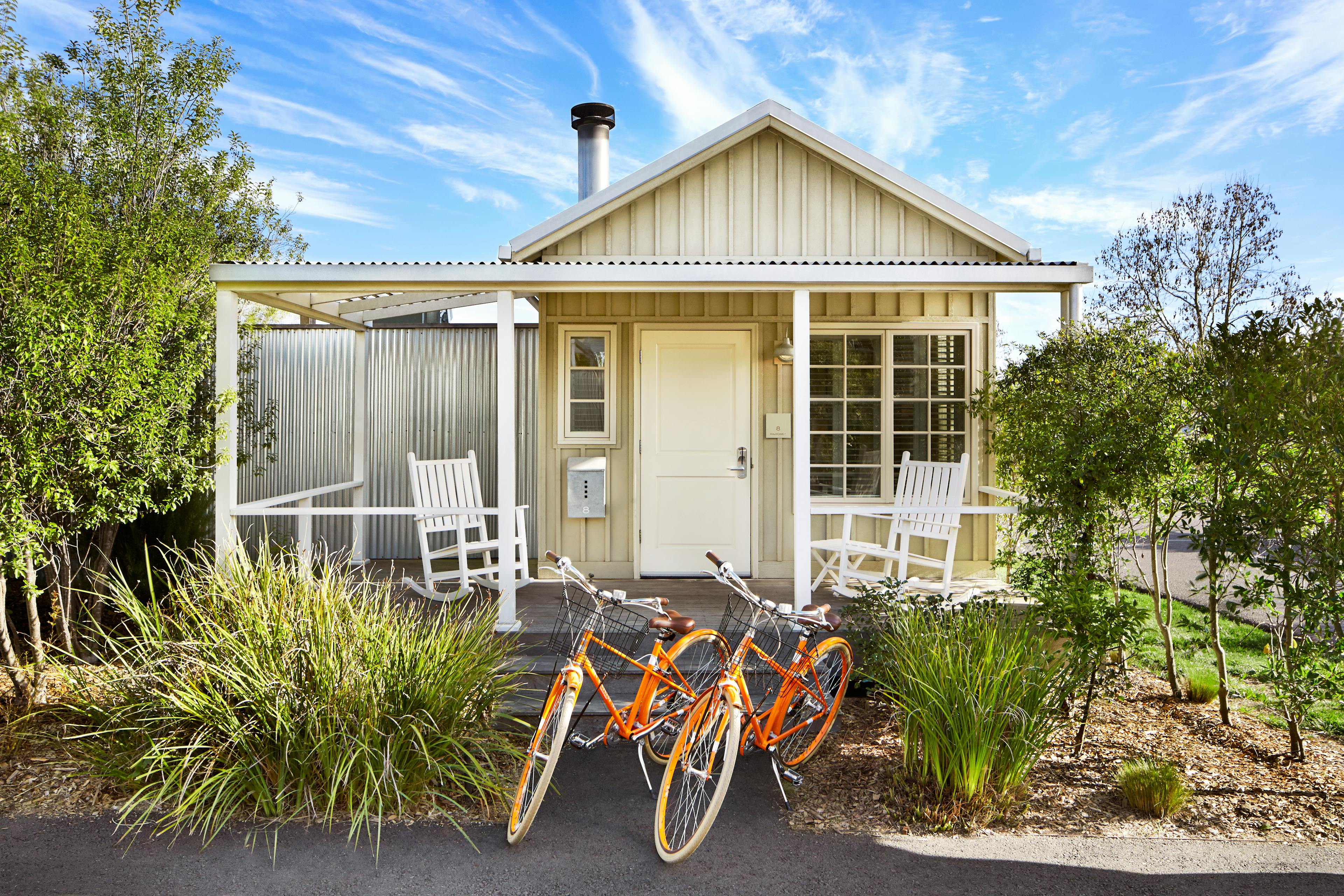 The exterior of a white cottage at the resort with a porch with two chairs and two orange bikes in front, surrounded by greenery.