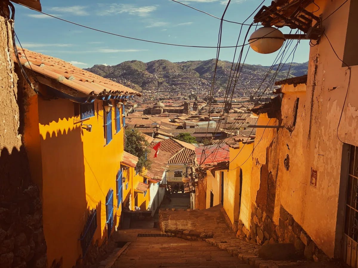 A quaint cobblestone street meanders down between traditional buildings, leading the eye to a bustling city cradled by mountains.