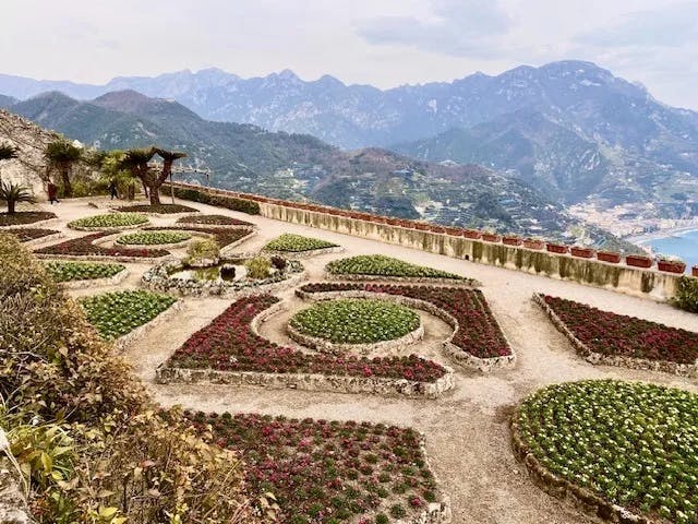 The image shows a terraced garden with patterned flower beds overlooking mountainous terrain and a body of water.