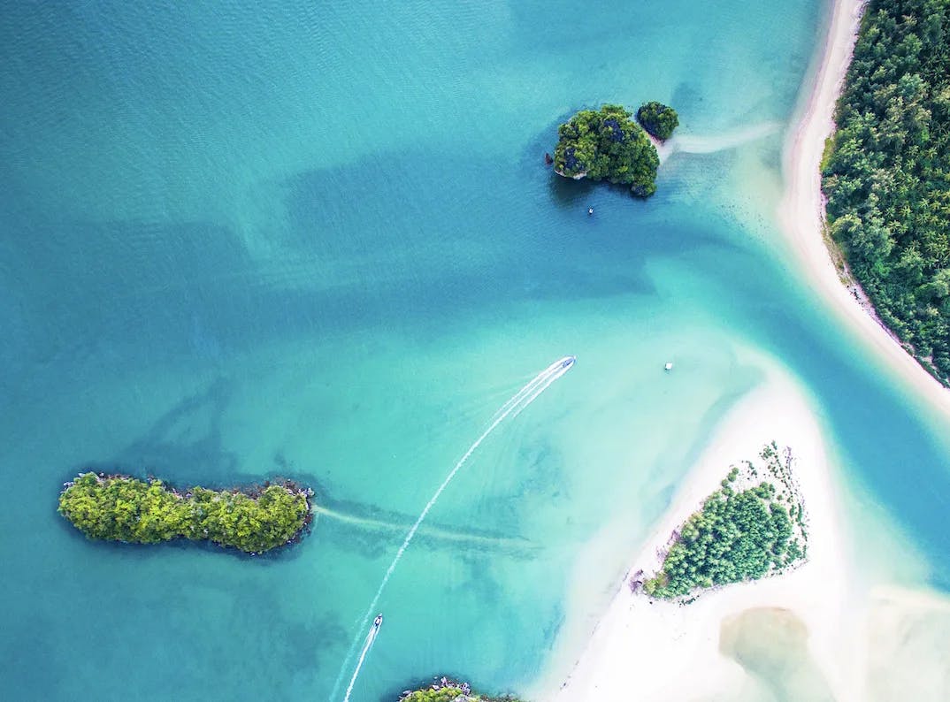 A birds-eye-view of a turquoise blue water, white sand beaches, sandbars, trees and a boat sailing across the water. 