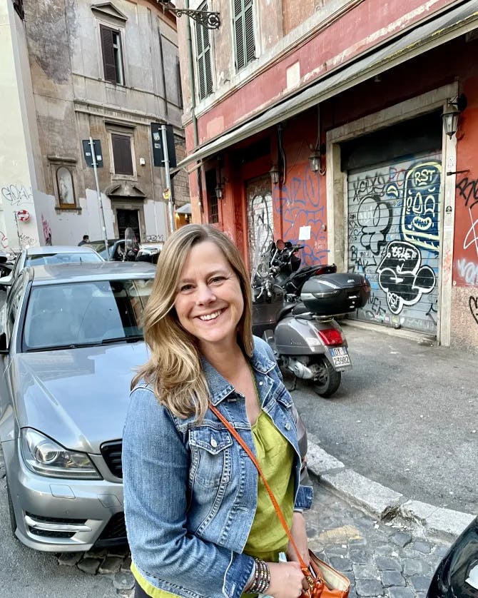 Jennifer wearing a green top and denim jacket standing outside on a city street in front of a car, motorcycle and red building covered in graffiti 