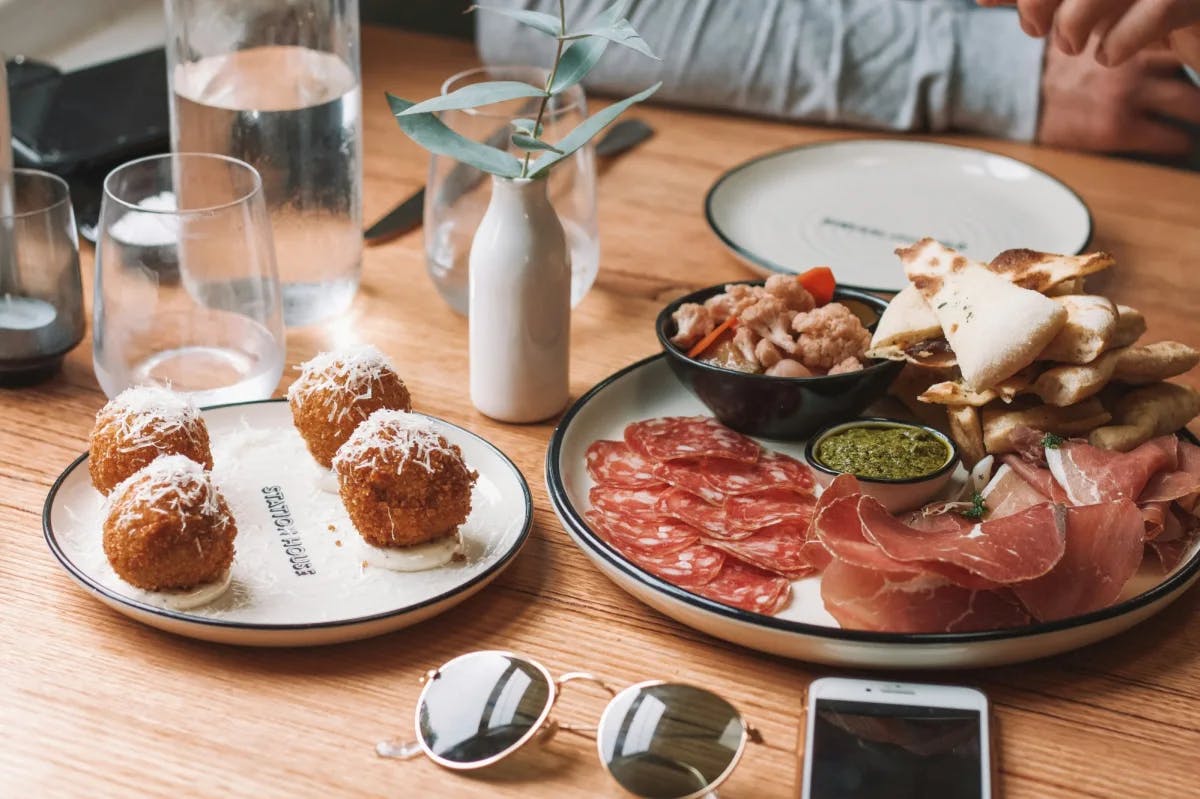 This image depicts a table of food consisting of arancini, cured meats, crackers, dips, plates and drinking glasses. 