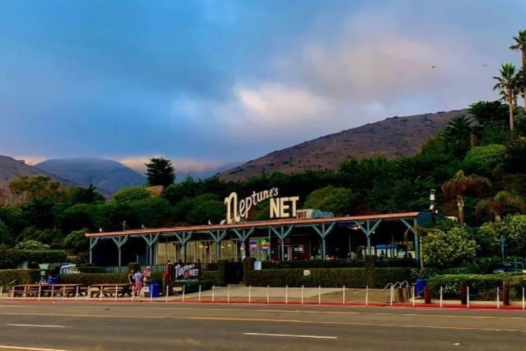 A photo of an outdoor restaurant with outdoor patio seating and a sign on top reading "Neptune's Net" with a mountain canyon in the background. 
