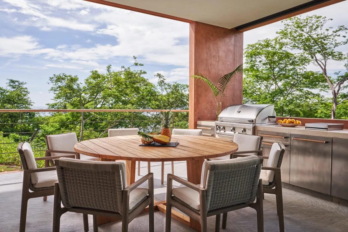 A full outdoor kitchen stands behind a circle table surrounded by comfy outdoor seating on a roofed patio