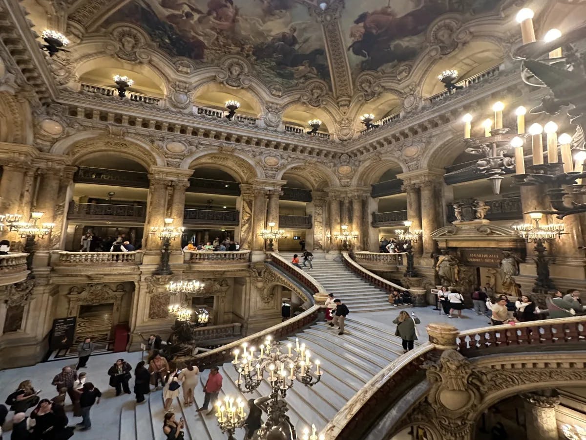 The ornate interior of the Paris Opera House with tourists walking on the grand staircase.