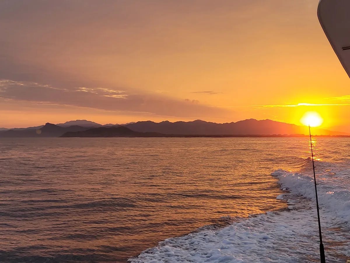 A view of the amber sunrise over the ocean and coastline, taken from the back of a fishing boat.