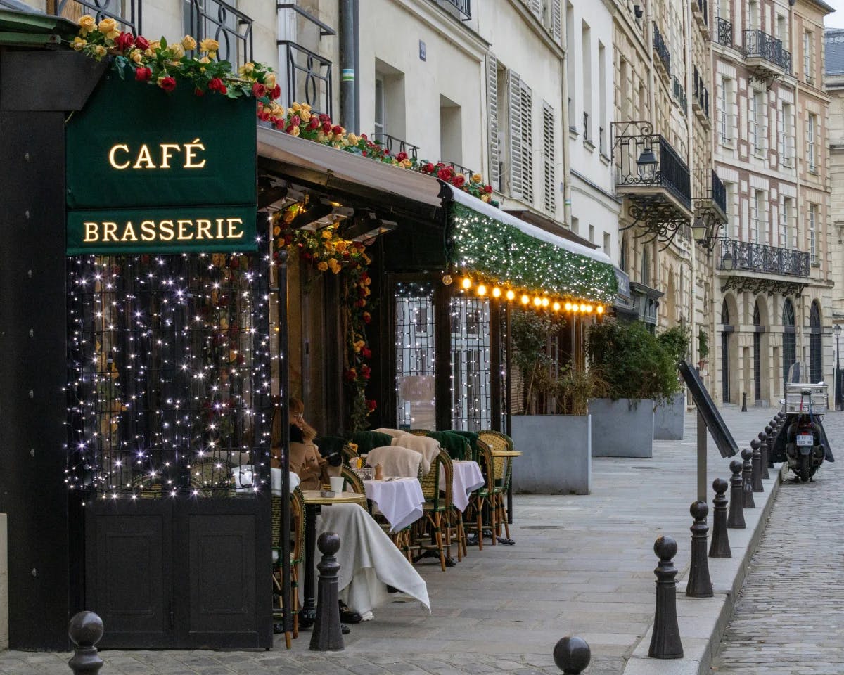 Outdoor seating in Paris with traditional French-style buildings, flowers on a balcony and a green awning that says "café / brasserie."