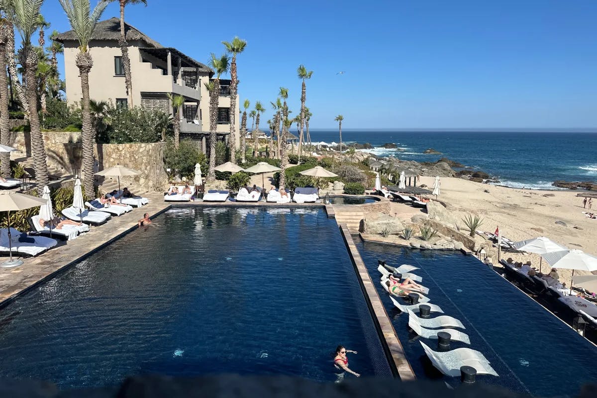 A blue pool surrounded by white lounge chairs and palm trees next to the beach with a beige building in the background.