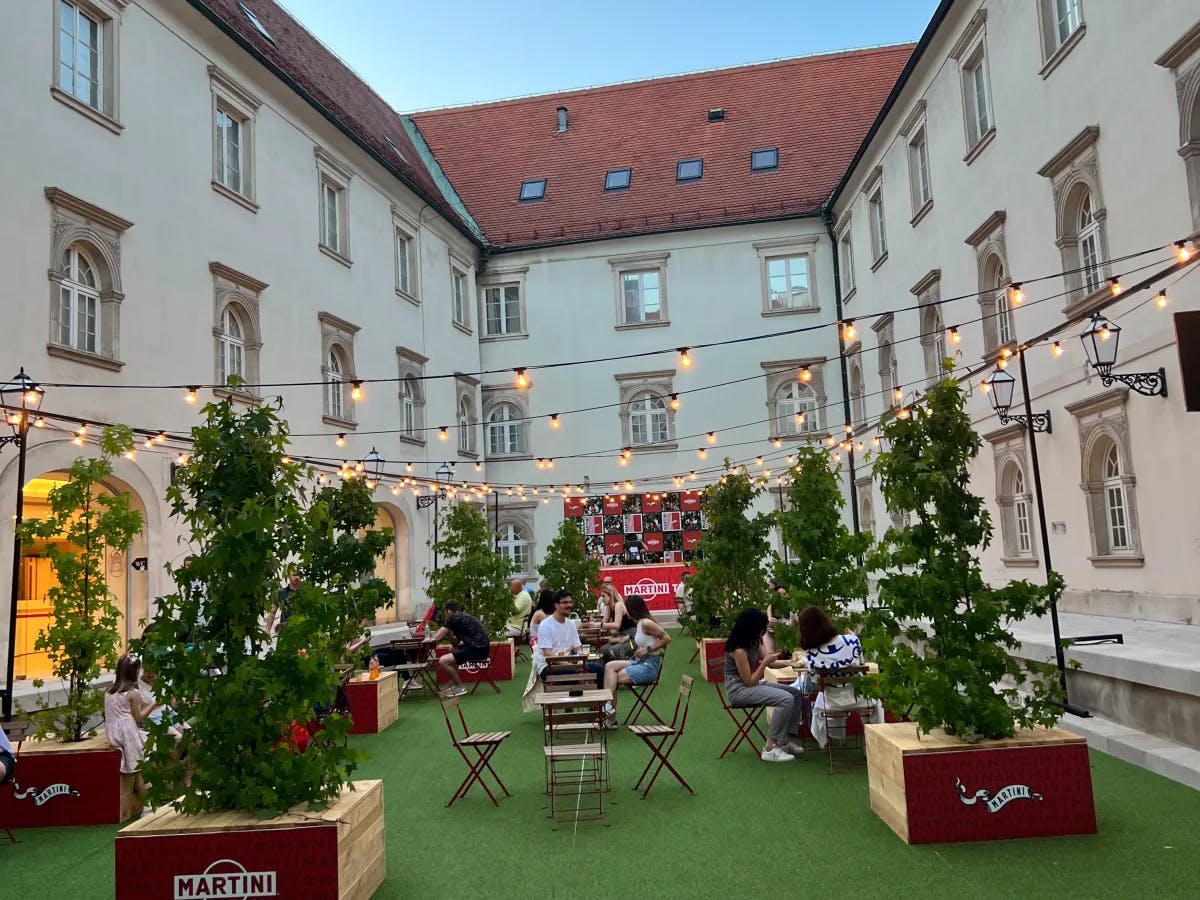 A cocktail garden with plants, string lights, bistro tables and chairs and a white building with red roofing.