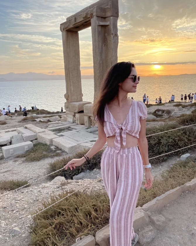 Alayna in a pink and white striped outfit posing in front of an archaeological structure by the sea at sunset