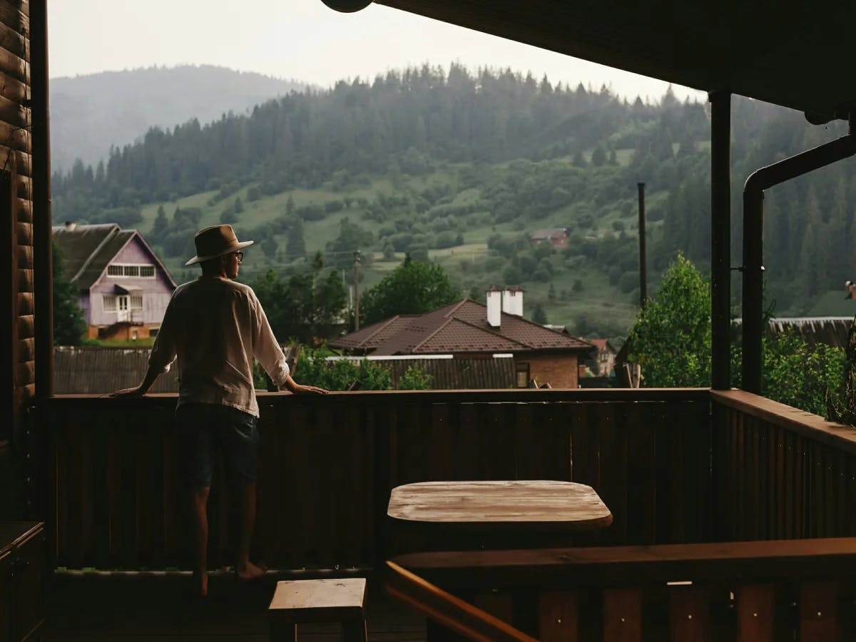 A person on a wooden porch, gazing at a serene rural setting with homes and forested hills.