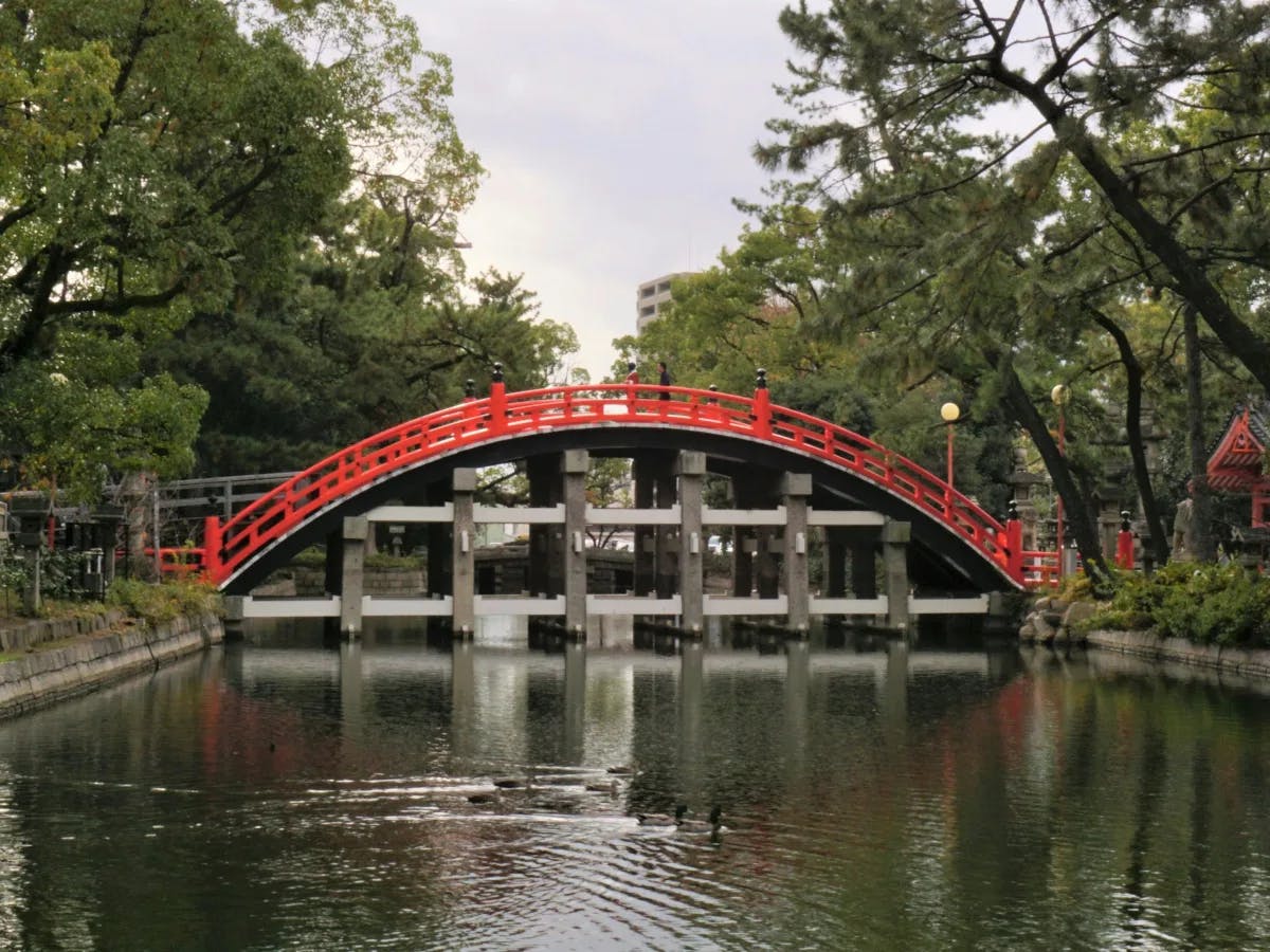 A red-arched bridge spans over a tranquil pond, surrounded by lush greenery and trees.