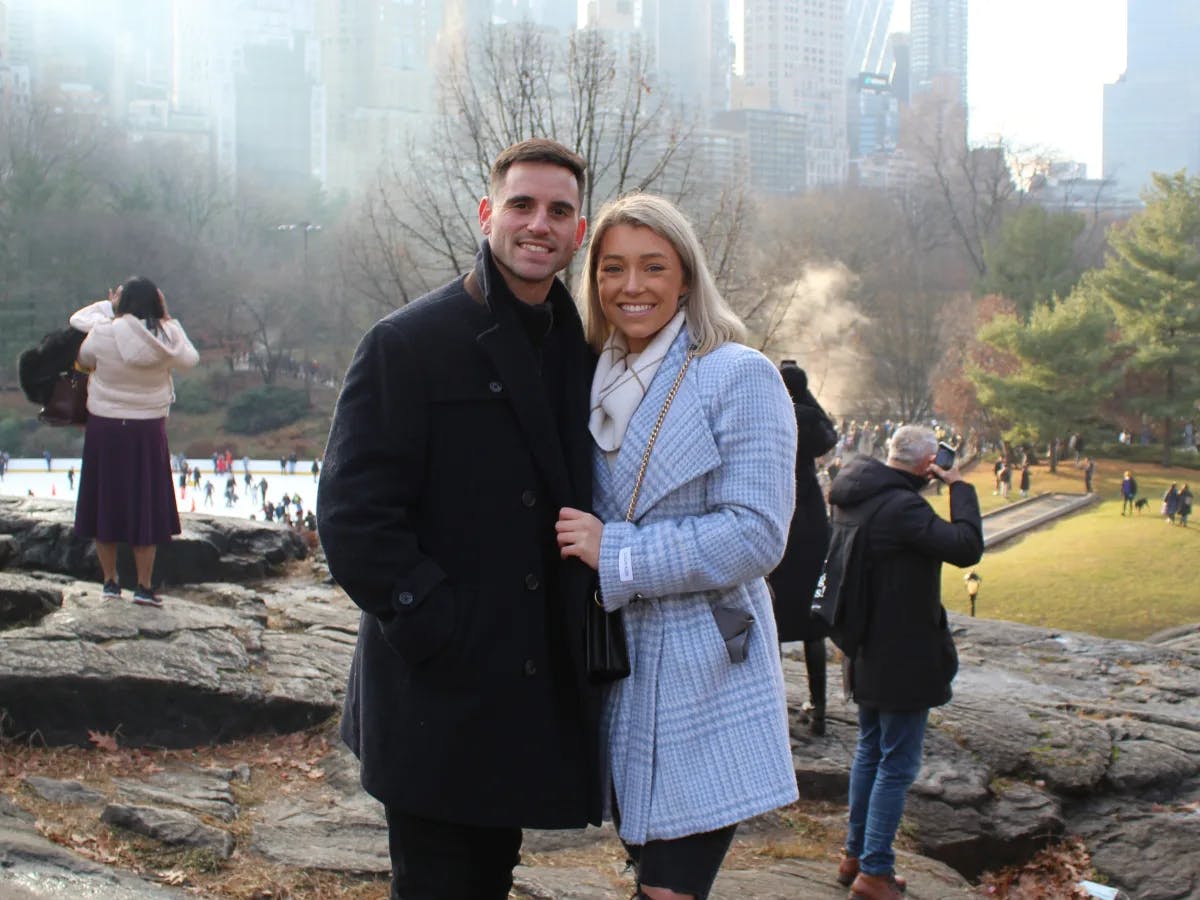 A couple posing for a photo in Central Park. There are people in the background, as well as trees, grass and tall buildings. 