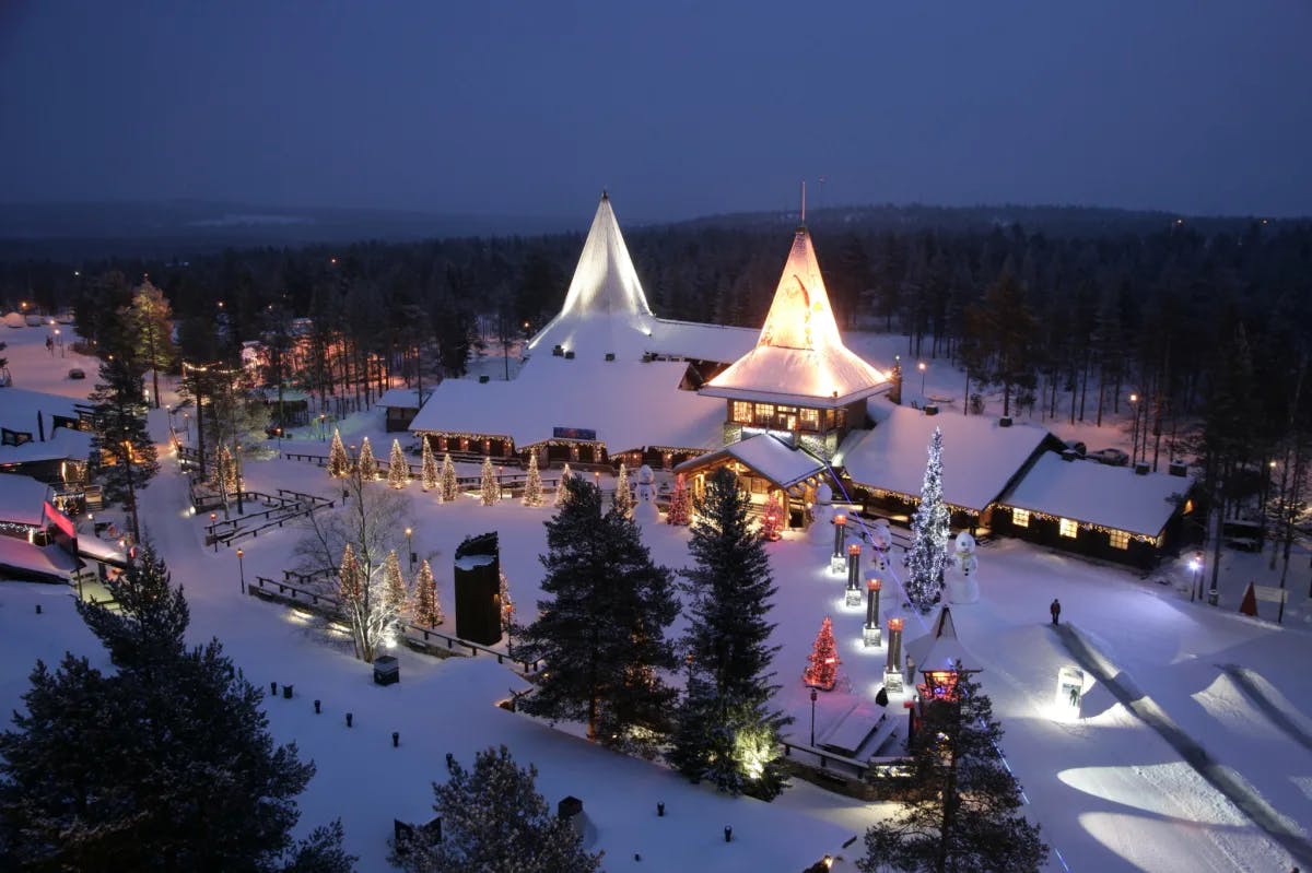 An aerial view of a lit-up village at night with a long white building with steepled roofs, Christmas trees and hills in the distance.