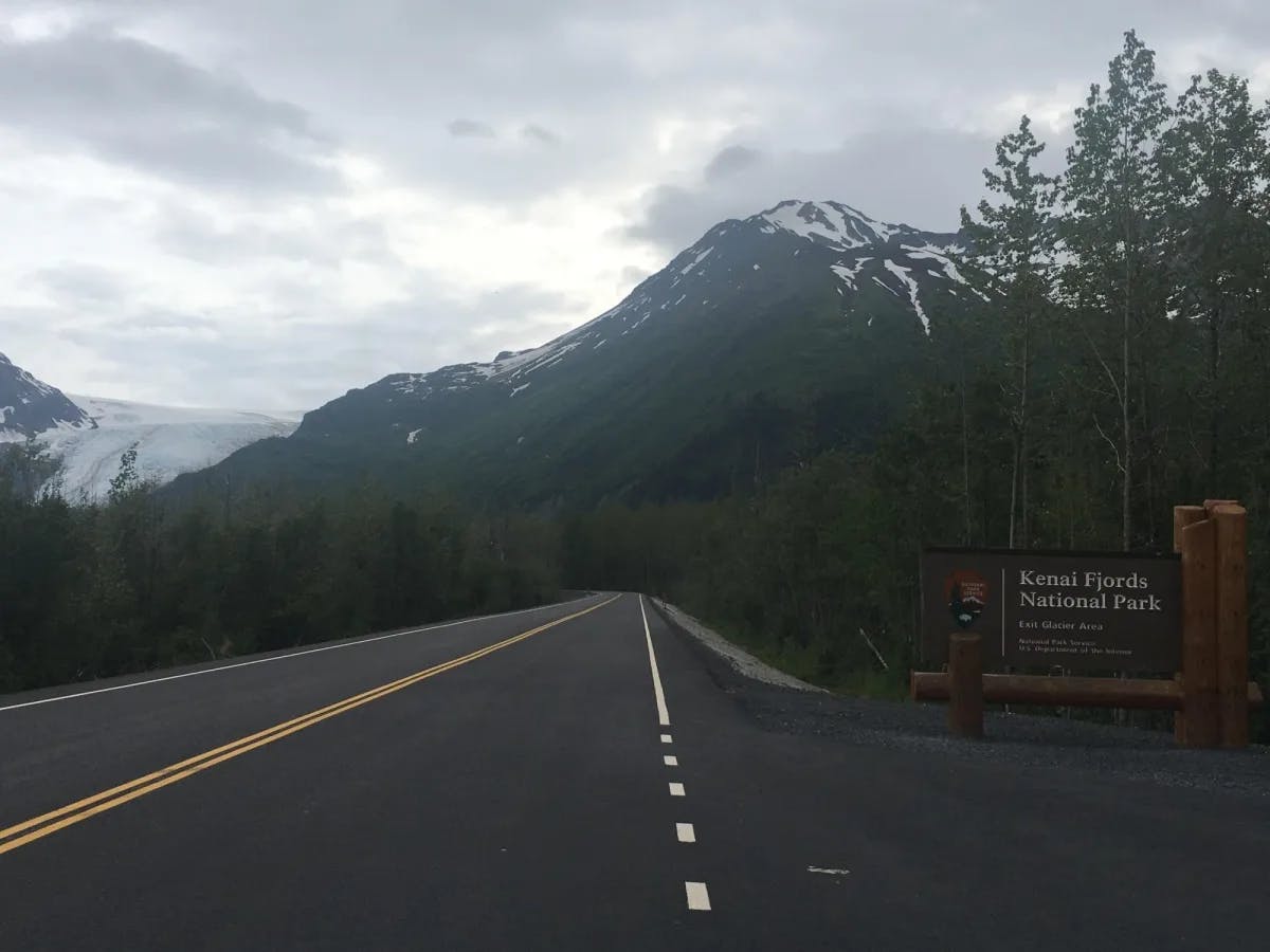 Road with snow covered mountains at the back and board saying Kenai Fjords National Park. 