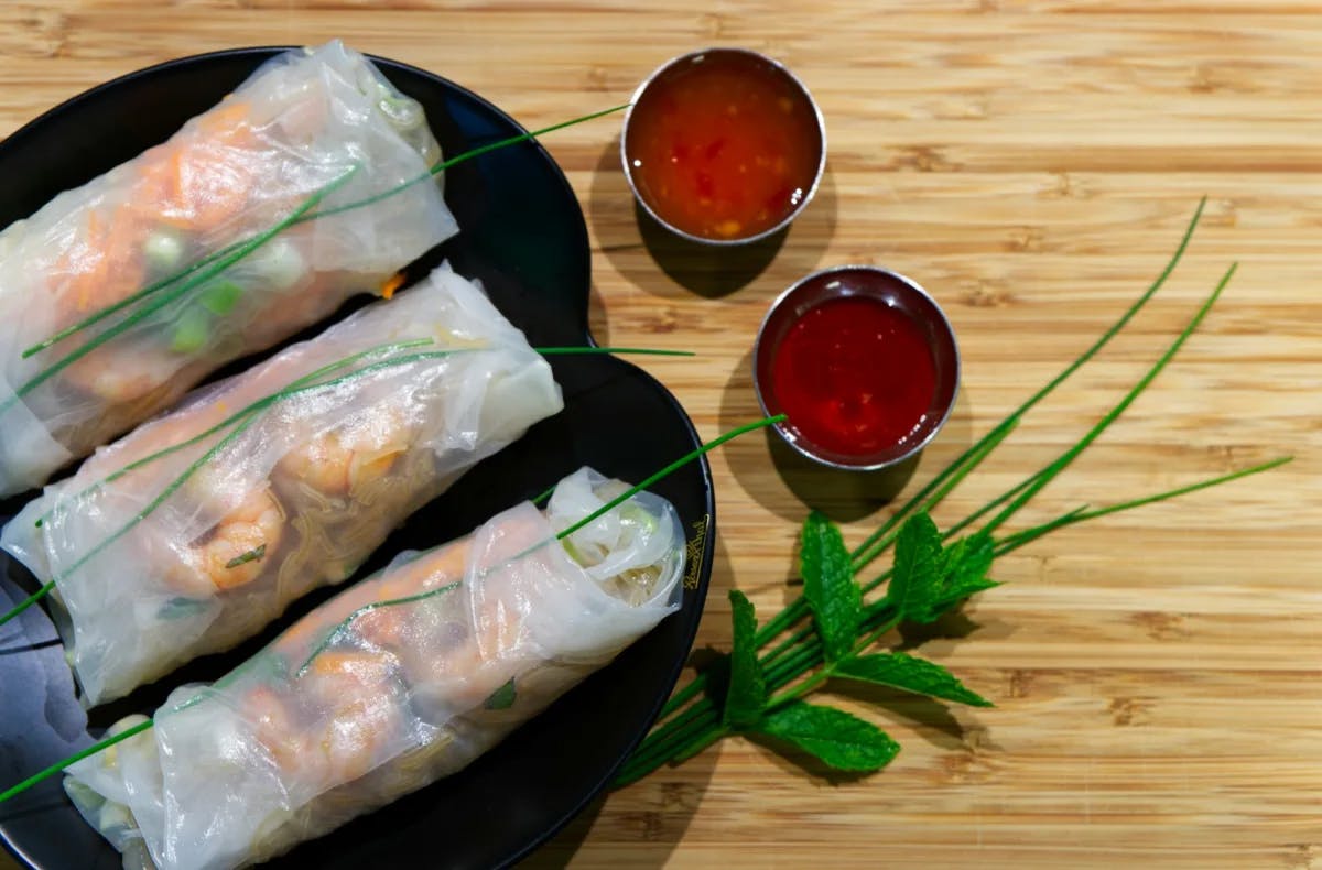 A plate of three freshly prepared spring rolls, topped with green herbs and two small portions of a red dipping sauce on the side.