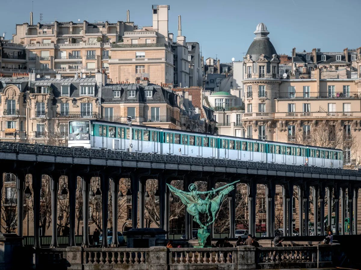 The image depicts a green metro train crossing a bridge adorned with sculptures in a bustling urban landscape.
