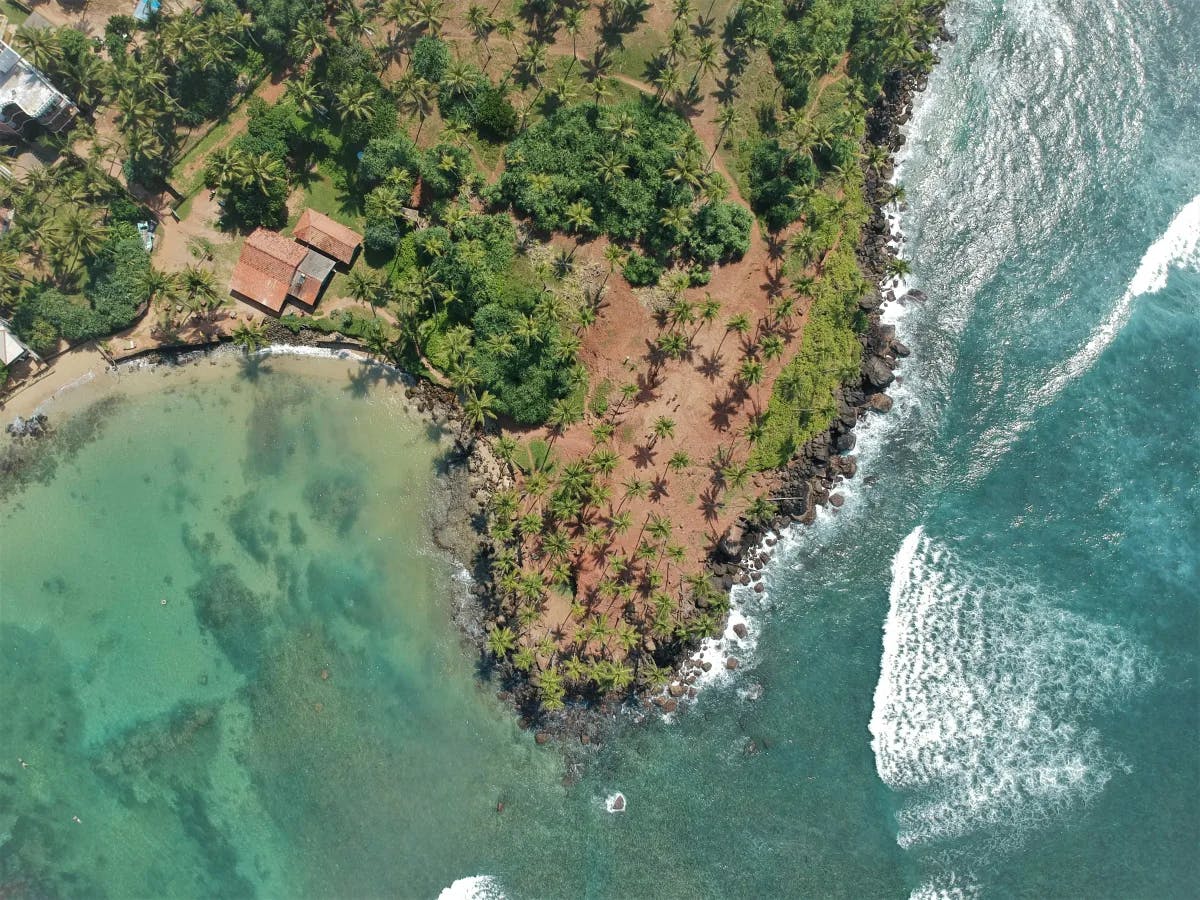 An aerial view shows a coastal landscape with a clear demarcation between a calm turquoise lagoon and rougher blue sea waves, bordered by lush greenery and a solitary building.