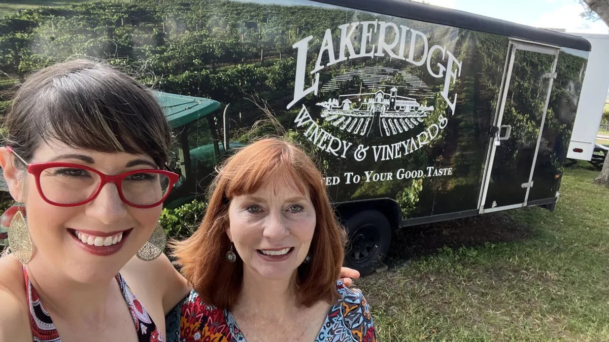 Two women posing for a selfie in front of a trailer with a sign that reads Lakeridge Winery.