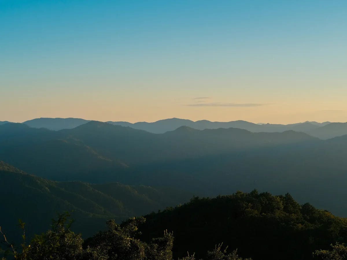 A serene landscape of layered mountain silhouettes under a gradient sky from blue to yellow, capturing nature's tranquil beauty at sunrise or sunset.