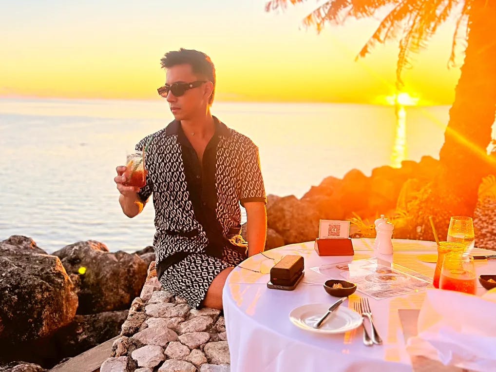 A man in black and white shirt, shorts sitting outdoors on beach with table in front with drinks. 