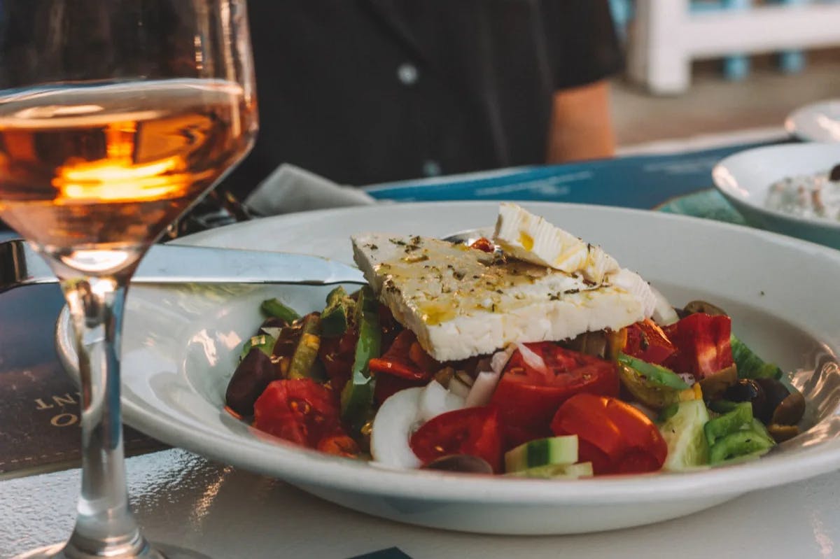 Greek salad on a white plate next to a glass of wine. 