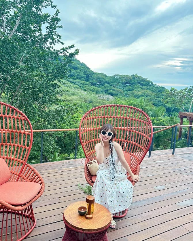 Jessica sitting on a red chair near a second red chair and small table atop a wooden deck that looks out over a tropical forest
