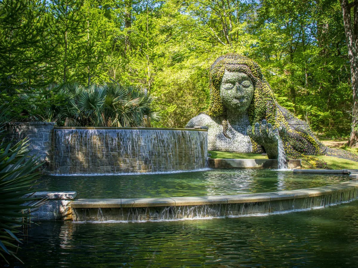 A picture of a water fountain with a stone woman sculpture on the right, in front of a lush assortment of trees. 