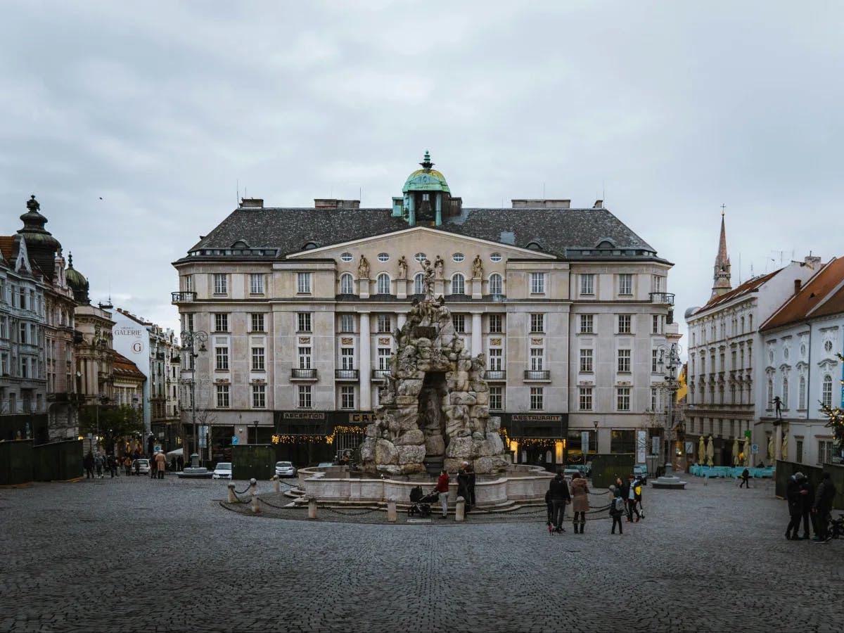 The image depicts a tranquil European square with classic architecture and a stone fountain, enlivened by people strolling around.