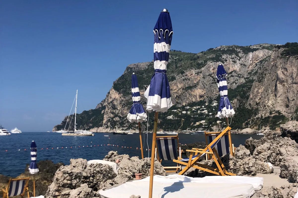 La Fontelina is an iconic beach club and restaurant located on the scenic coastline of Capri. Picture is of blue and white umbrellas on a beach with rocky outcroppings in the distance.