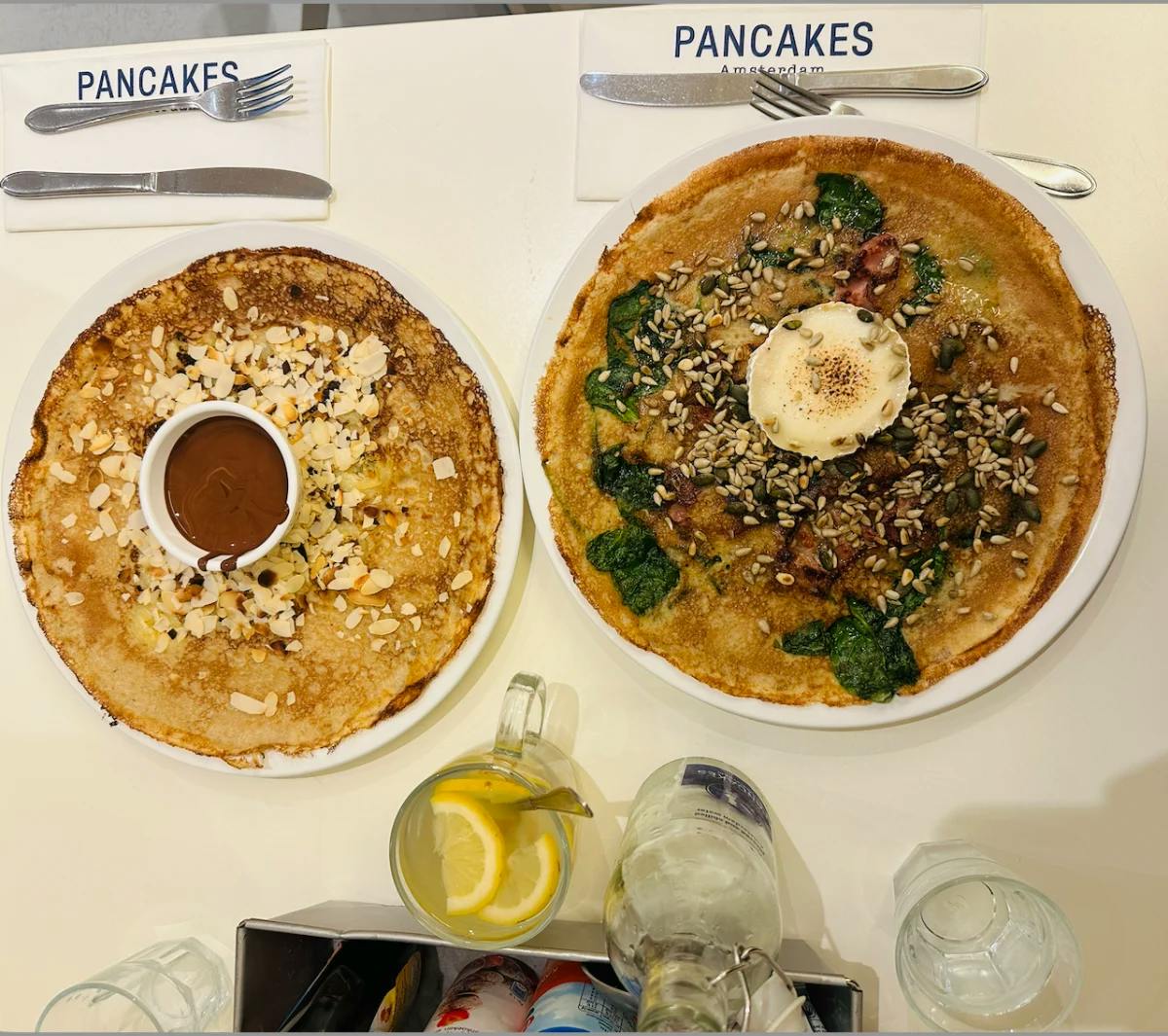 Two pancakes, one with spinach and seeds and the other with almond shavings and Nutella, on a table with cutlery. 