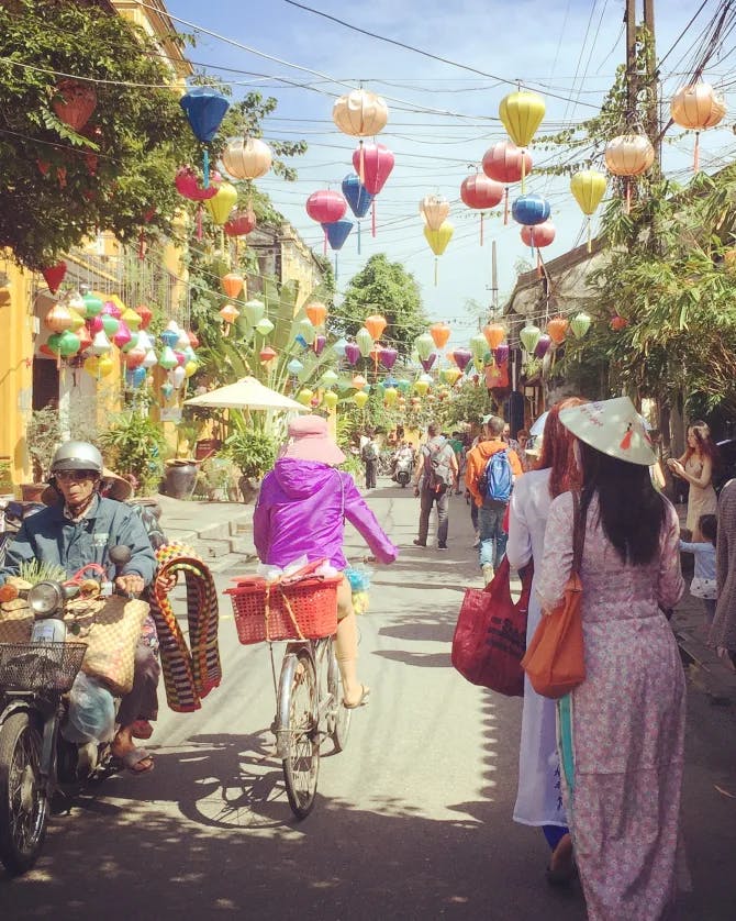 A view of a busy street with people on bicycles and colorful lanterns lining the street on a sunny day.