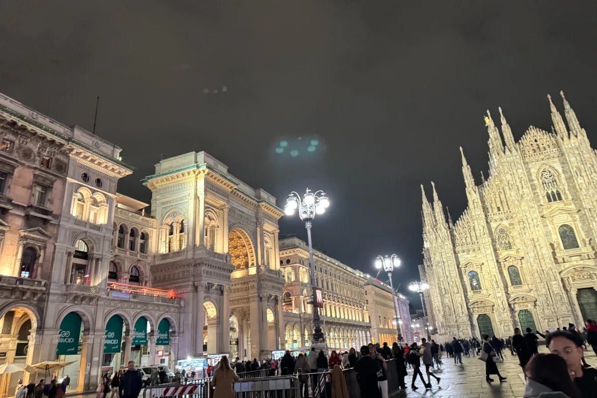 A low-angled shot of streets surrounded by European buildings during the night. There are people walking around in the forefront beneath city lights. 