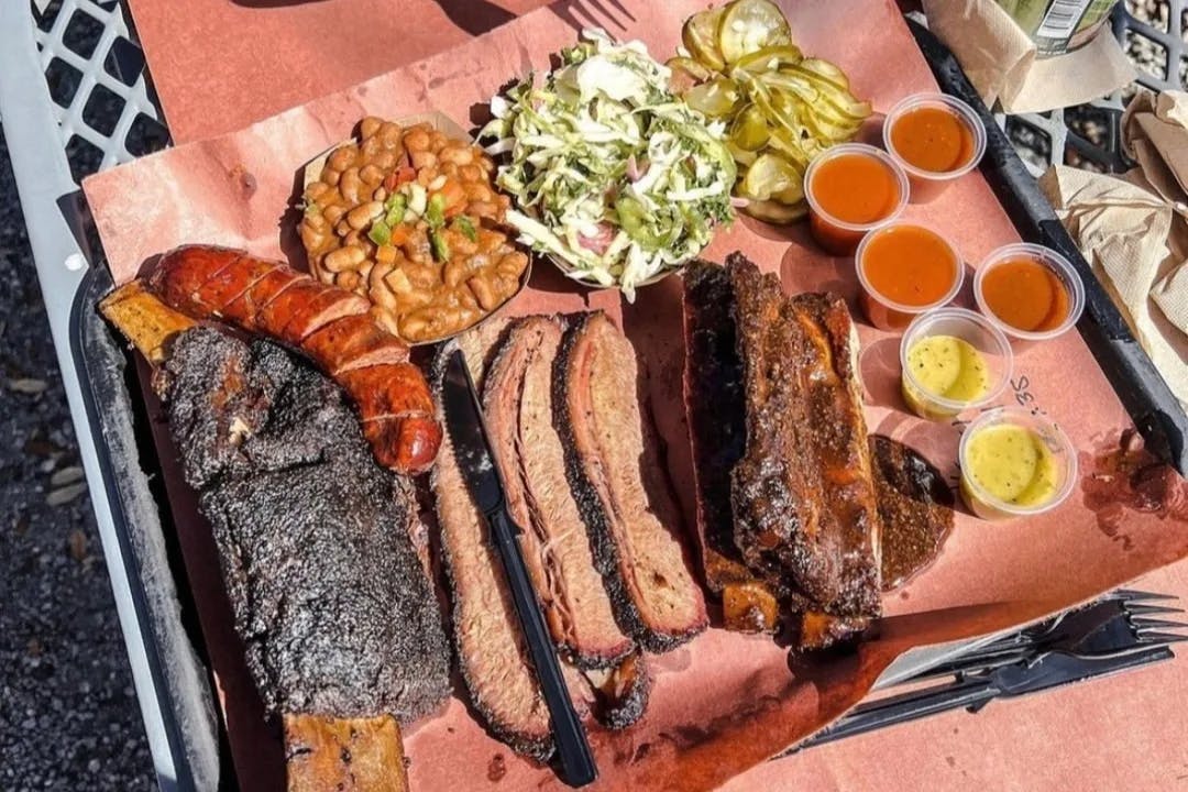 A platter of smoked meats and cups of various sides on a red tablecloth 