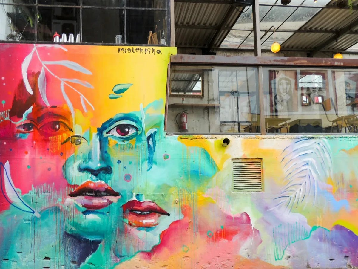 An abstract street art mural of a woman's face with various colors. There is a warehouse-style building in the upper right hand corner. 