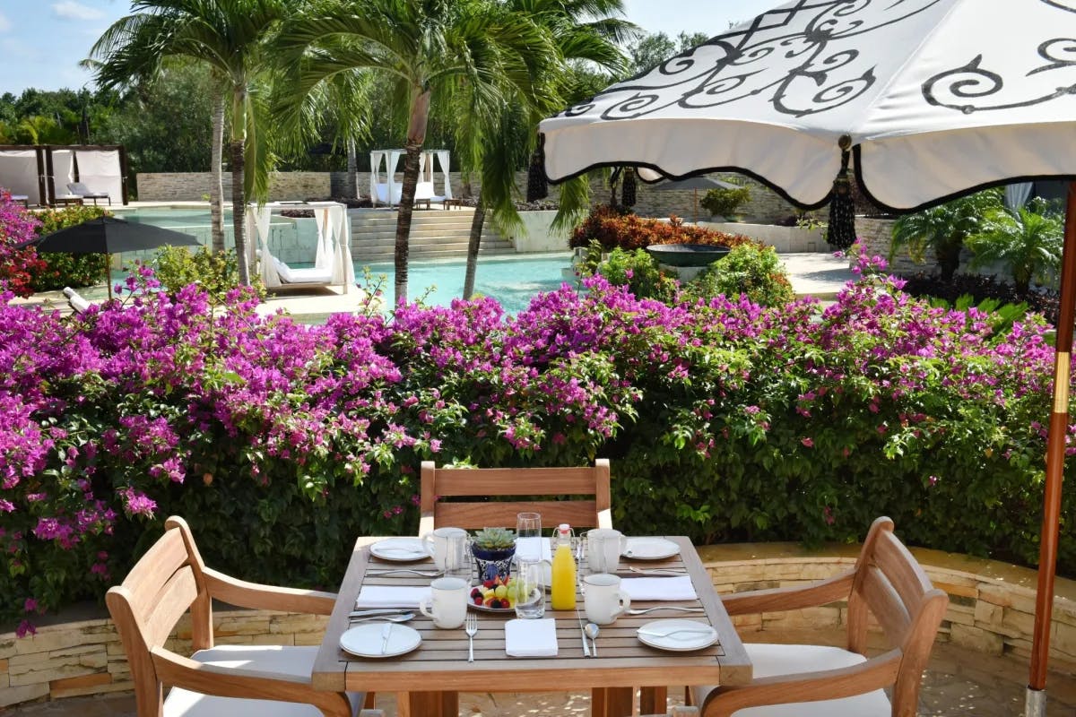 outdoor dining table with purple flowering bush and umbrella and pool in the background