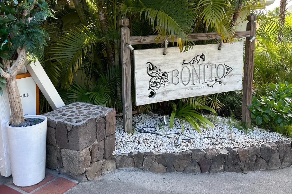 The entrance to Bonito, a French restaurant in St. Barth with a wooden sign surrounded by lush foliage on a sunny day.