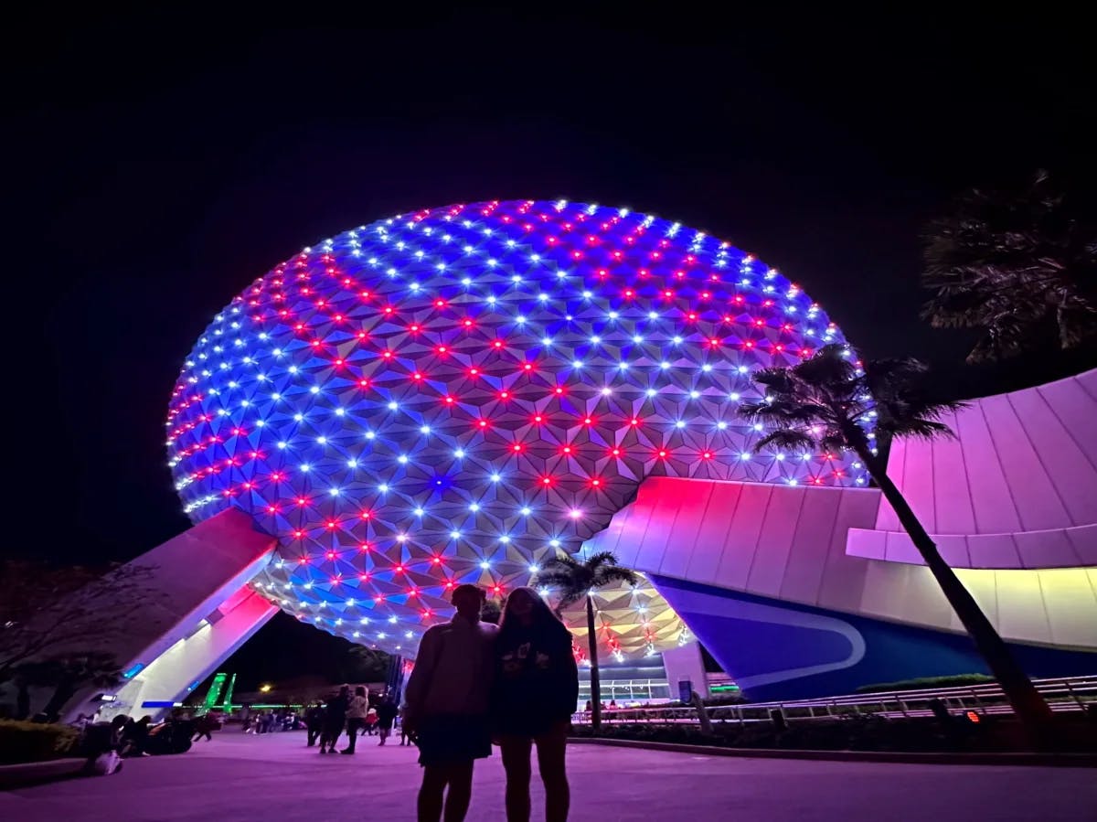 Two people standing in front of a lit-up multi-colored round-shaped building during nighttime.