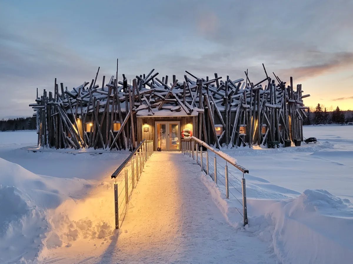 Arctic Bath, a lit-up hut with sticks on the exterior at the end of a snowy pathway.