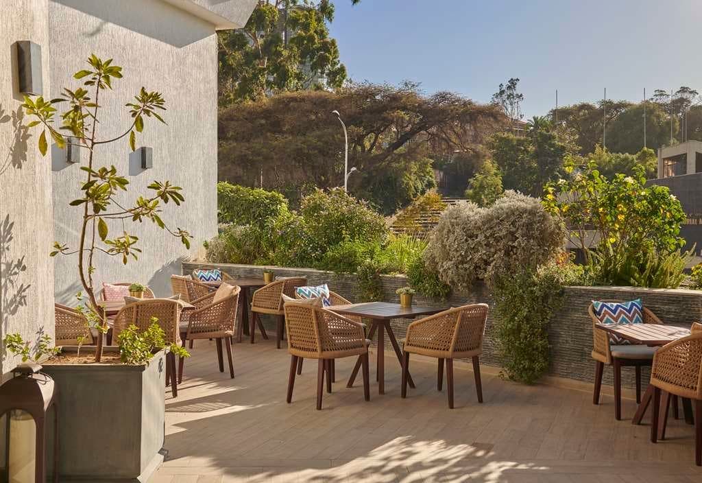 Outside sitting area with patio seating surrounded by lush greenery on a sunny day. 