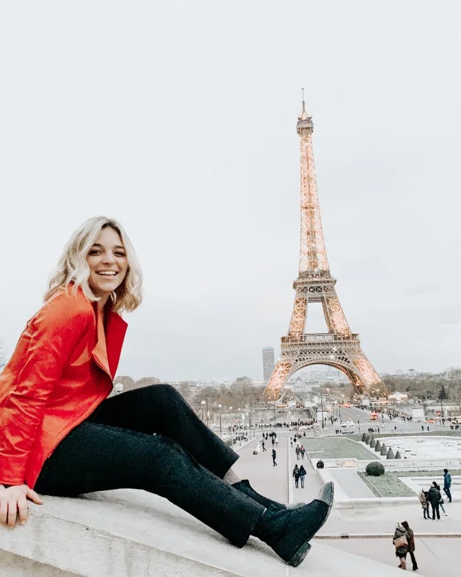 Hannah in bright red jacket sitting on a ledge with a beautiful view of the Eiffel Tower behind her